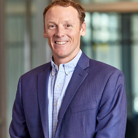 A man with short, light brown hair smiles while wearing a blue suit jacket over a light blue checkered shirt, standing indoors in front of a blurred office background.