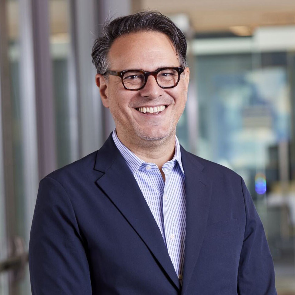 A man wearing glasses and a navy blazer over a light blue striped shirt smiles while standing indoors in front of blurred glass walls.