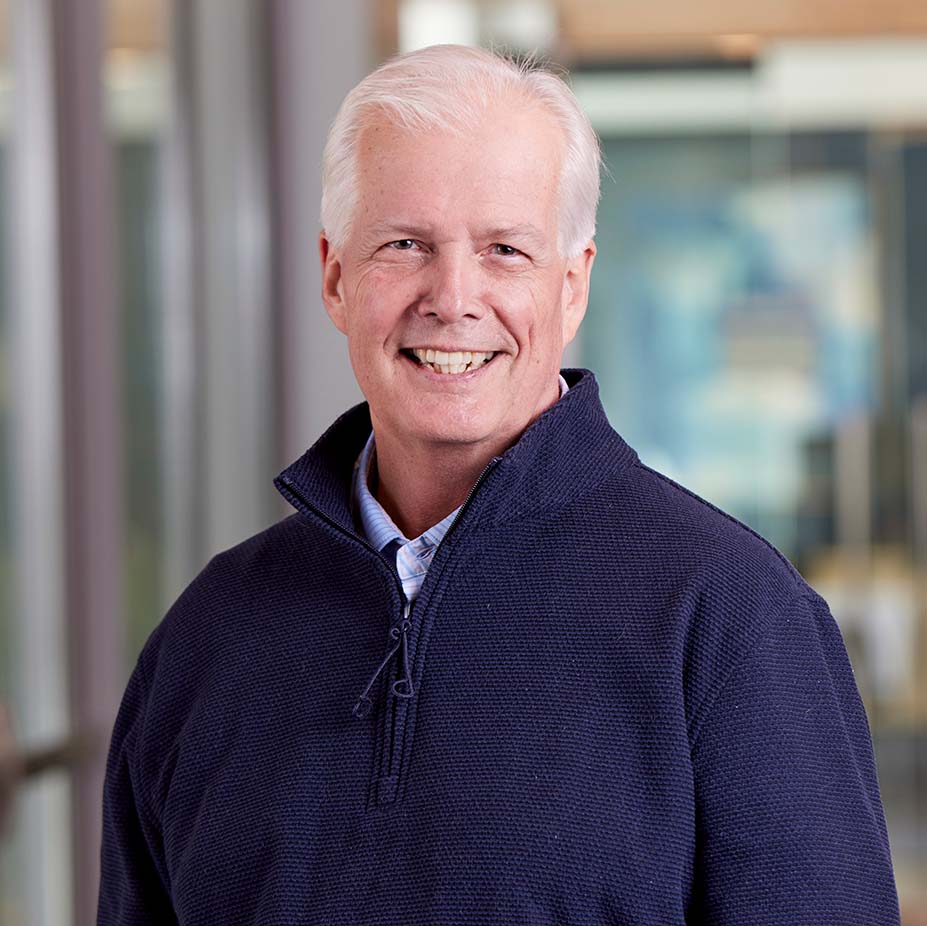 A smiling older man with short white hair wearing a navy blue pullover stands indoors, in front of a blurred background with large windows and soft natural light.