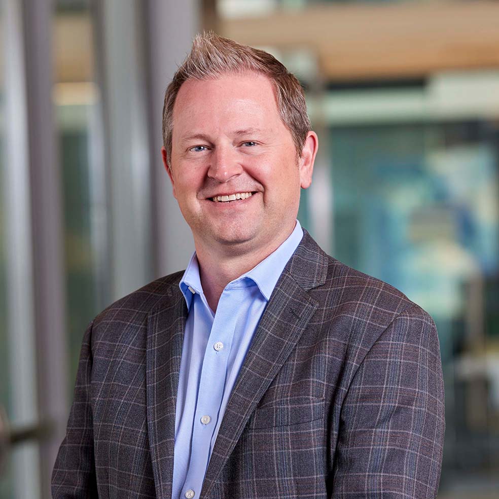 A smiling man with short, light hair wears a blue shirt and a gray plaid suit jacket, standing indoors in front of a blurred office background.