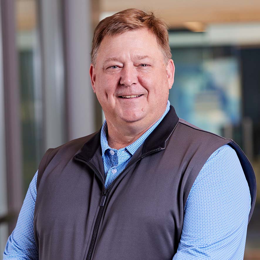 A middle-aged man with light brown hair smiles at the camera. He is wearing a light blue shirt and a gray vest. The background is indoors with blurred office elements.