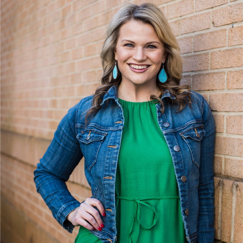A woman with wavy blonde hair smiles while standing against a brick wall. She is wearing a blue denim jacket over a green dress and turquoise earrings, with one hand on her hip.