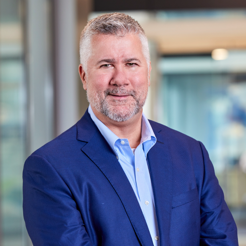 A middle-aged man with short gray hair and a beard, wearing a blue suit jacket and light blue shirt, stands smiling indoors in a modern office setting with blurred background.